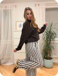 Woman in a black top and striped pants standing in a room with curtains and a plant.