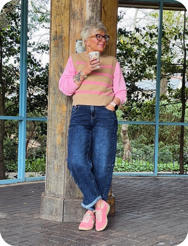 Woman in pink sweater and blue jeans standing next to a wooden post with trees in the background